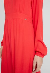 Robe rouge vif avec manches longues, corsage ajusté et tissu texturé. Une petite étiquette de marque est attachée à la couture de la taille.