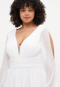 Weißes Kleid mit langen, transparenten Ärmeln, tiefem V-Ausschnitt und geraffter Taille. Der Stoff ist leicht und hat eine fließende Silhouette.