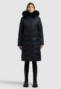Marineblaue lange Parka mit Kapuze aus Kunstfell, Reißverschluss vorne, zwei Seitentaschen und glattem, glänzendem Finish. Getragen mit schwarzen Leggings und Stiefeln.