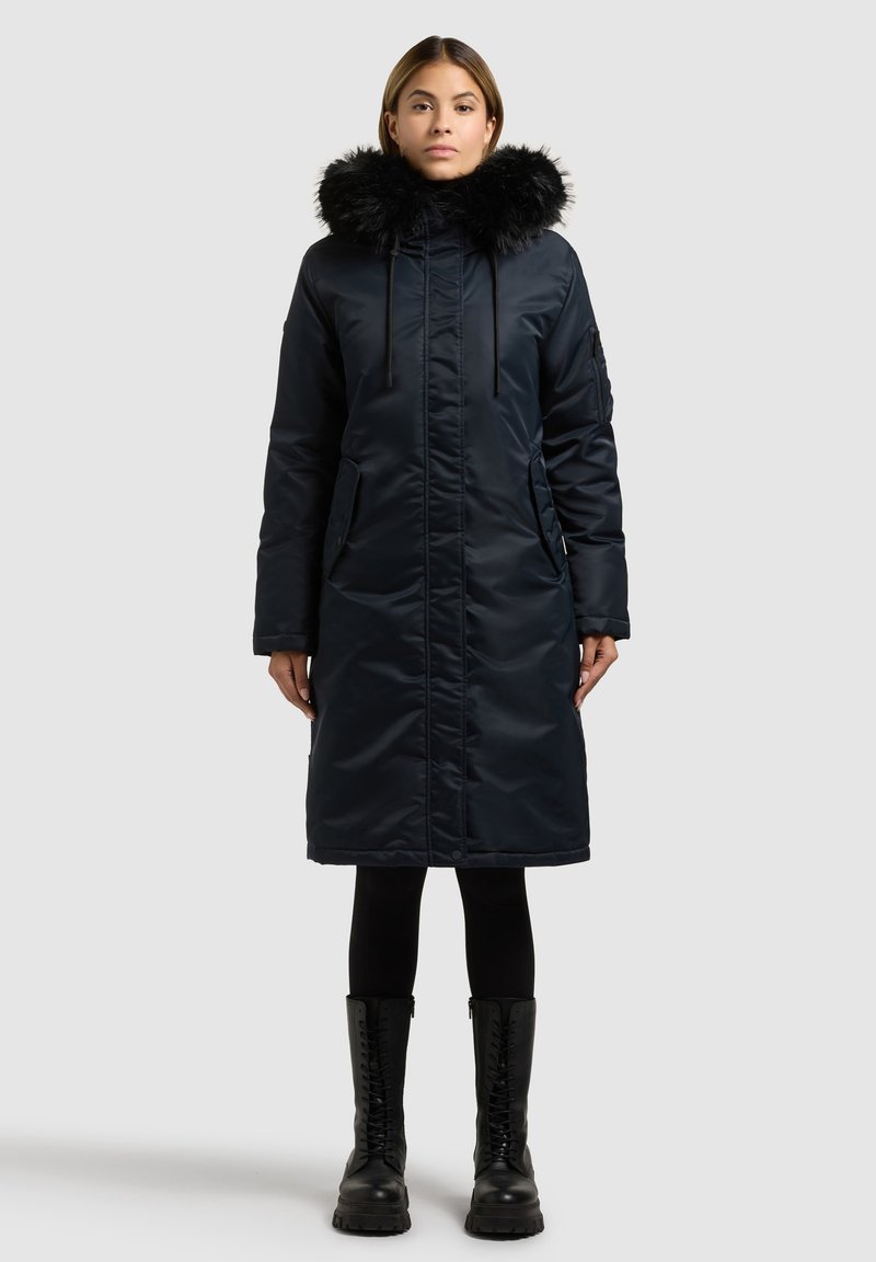 Parka lunga blu scuro con cappuccio in pelliccia sintetica, cerniera frontale, due tasche laterali e finitura liscia e lucida. Indossata con leggings neri e stivali.