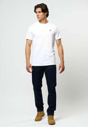 Weißes T-Shirt mit kurzen Ärmeln und einem kleinen schwarzen Logo auf der linken Brust, kombiniert mit dunkelblauer Jeans und hellbraunen Schnürstiefeln.