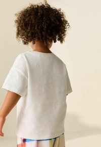 Weißes Leinen-T-Shirt mit lässiger Passform, kurzen Ärmeln und rundem Ausschnitt, kombiniert mit bunten, gemusterten Shorts.