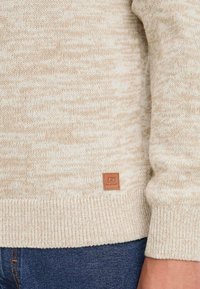 Suéter de punto beige con ribete en el dobladillo y los puños, que se lleva sobre unos jeans de mezclilla azules, con un pequeño parche de cuero marrón de la marca cerca de la cintura.