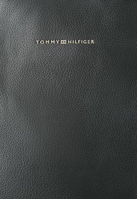 Svart lærmateriale med en strukturert overflate. En gullfarget "TOMMY HILFIGER"-logo er plassert midt på toppen.