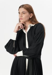 A model with long, straight hair thoughtfully poses, wearing a pleated black dress with a white collar and cuffs, against a neutral background.