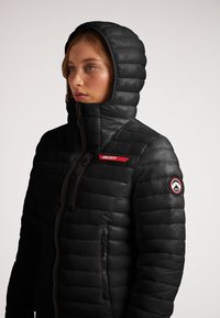 Giubbotto imbottito nero con colletto alto e cappuccio, dotato di una design a righe orizzontali, tasche con zip e un accento del logo rosso sulla parte frontale.