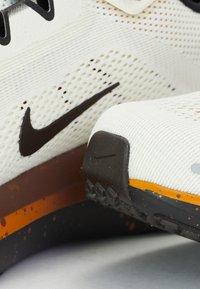 Nike running shoe featuring a white mesh upper with a black swoosh, orange and brown speckled sole, and textured rubber tread for grip.