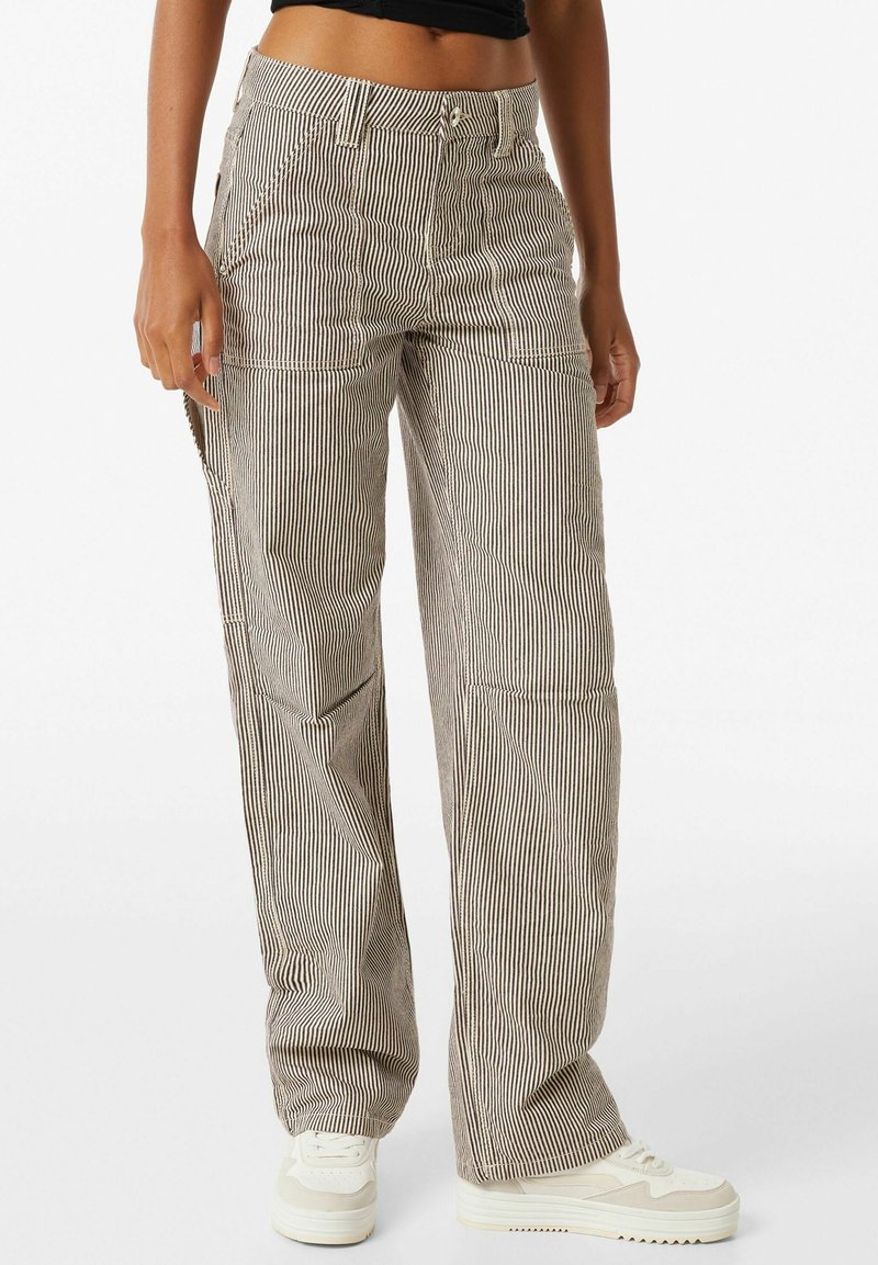 Femme portant un pantalon à rayures verticales beige et noir et des baskets blanches à plateforme, debout devant un fond blanc uni.