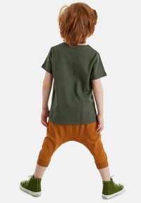 Camiseta de algodón verde, pantalones naranjas holgados con tiro caído diseñado y zapatillas verdes altas con suelas blancas. Vista trasera.