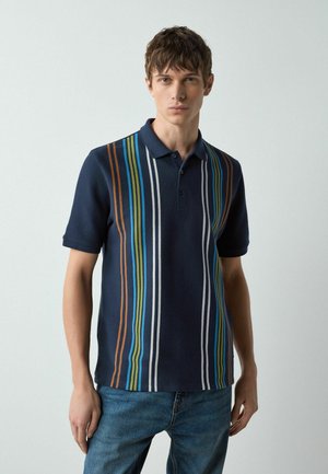 Young man wearing a navy blue polo shirt with vertical multicolor stripes and blue jeans, standing against a plain light background.