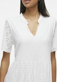 Robe en coton blanche avec des manches courtes, présentant un décolleté en V, un motif de broderie à œillets et une jupe évasée. Détail de tissu texturé visible.