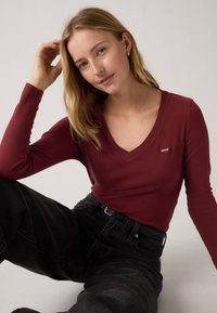 Bordeaux langermet V-hals topp laget av mykt stoff, kombinert med høyt liv svarte jeans, med belte og en liten logo detaljer.