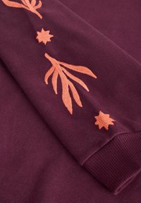 Bordová mikina s vyšívanými koralovými listami a hviezdami na rukáve. Mäkká textúra s rebrovanými manžetami.