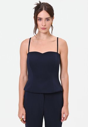 Donna con capelli castani raccolti in uno chignon basso indossa un top nero aderente senza maniche con sottili spalline e pantaloni neri abbinati su sfondo semplice.