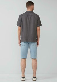 Kurzärmliges, kragenloses Hemd aus grauem Stoff mit feinem Muster, kombiniert mit hellblauen Jeansshorts und Sneakers.