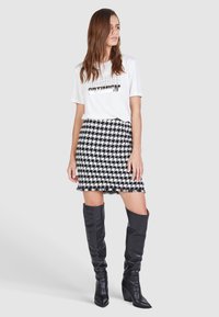 Weißes T-Shirt mit metallic-Text, kombiniert mit einem schwarz-weißen Hahnentrittmuster-Minirock. Das Model trägt kniehohe schwarze Lederstiefel.