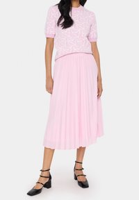 Ljusrosa stickad topp med abstrakt vitt mönster, parat med en plisserad rosa midikjol. Svarta blockklackskor fulländar outfiten.