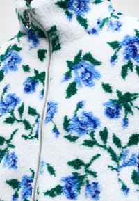 Veste en polaire blanche avec une fermeture éclair argentée et un motif floral bleu avec des feuilles vertes sur un tissu texturé.