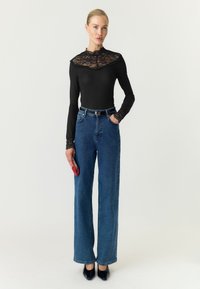 Femme portant un haut en dentelle noire à manches longues, jean bleu taille haute à jambes larges, talons noirs, tenant une petite pochette rouge sur un fond uni.