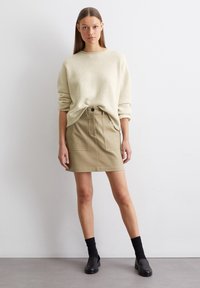 Pull léger beige avec une coupe décontractée et un col rond ; associé à une jupe mini kaki avec poches avant et fermeture à bouton. Chaussettes noires à la cheville et ballerines.