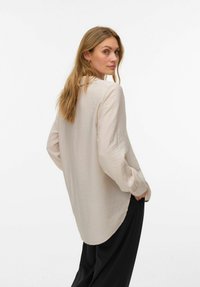 Hele varrukaga, helebeeži värvusega tekstuurse viimistlusega särk, ümarate äärete ja nööpidega varrukakätistega, mida on kombineeritud musta laia säärega pükstega.