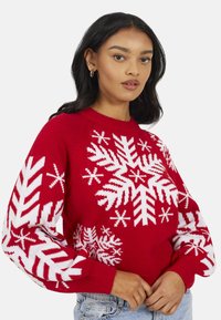 Pull tricoté rouge avec motif flocon de neige blanc et manches longues. Encolure ronde avec poignets et ourlet côtelés. Coupe décontractée, texture douce.