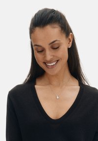 Sølvhalskæde med vedhæng, båret af en person i en sort V-hals sweater. Øreringene matcher halskæden, begge med et enkelt design.