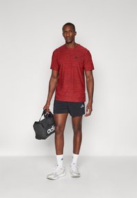 T-shirt de sport rouge à manches courtes avec un motif texturé et un logo noir, associé à un short noir et des chaussures de sport blanches, tenant un sac de sport noir.