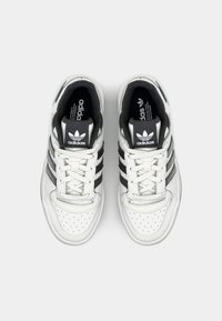 Bílé kožené tenisky se třemi černými pruhy, perforovanou špičkou, tkaničkami a polstrovaným límcem; na jazyku je logo Adidas.