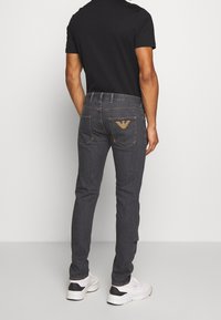 Dark denim jeans with a fitted design, featuring a subtle logo in tan on the back pockets, paired with a black t-shirt and white sneakers.