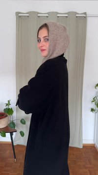 Zwarte buitenjas met lange mouwen, gecombineerd met een lichtbeige wollen hoofddoek. Op de achtergrond een lichte gordijn en een potplant.