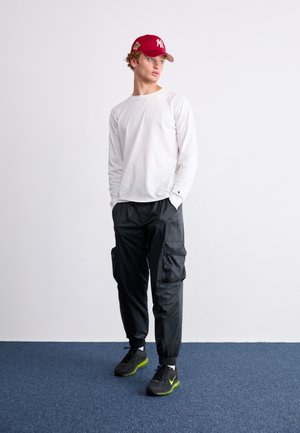 Young man standing with hands in pockets wearing red baseball cap, white long-sleeve shirt, black cargo pants, and black sneakers with green soles.