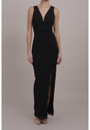 Femme portant une robe de soirée noire sans manches avec un décolleté en V profond et une fente haute sur la cuisse, associée à des escarpins beiges à bout pointu.