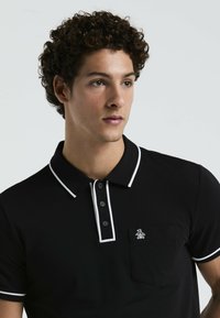 Schwarzes Polohemd mit weißen Akzenten, Knopfleiste mit drei Knöpfen und aufgesetzter Tasche. Verfügt über eine weiche Textur und ein gesticktes Logo auf der Brust.