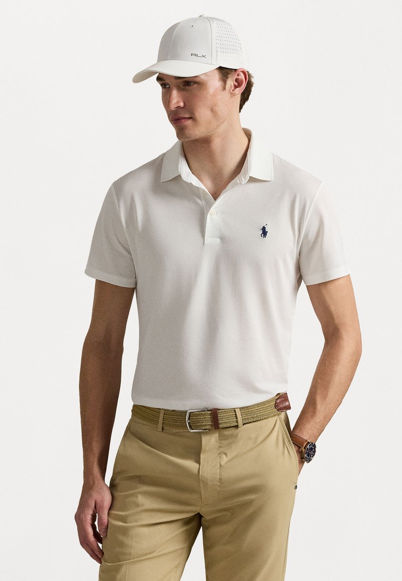 Camisa polo branca com colarinho, mangas curtas e um pequeno logótipo azul-marinho. Combinada com calças caqui e um cinto trançado. Boné branco.