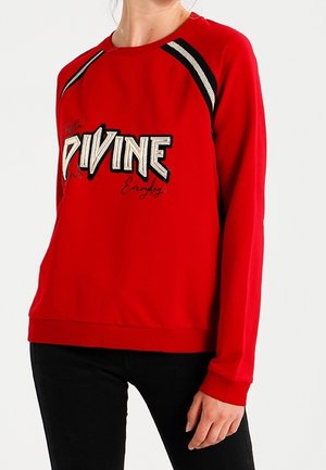 Femme portant un sweat-shirt rouge avec des rayures noires et blanches sur les épaules et le mot "DIVINE" en grandes lettres sur la poitrine.