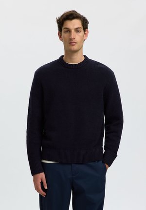Jeune homme aux cheveux courts et bruns portant un pull en maille bleu marine foncé et un pantalon bleu marine, debout avec une main dans la poche devant un fond uni.
