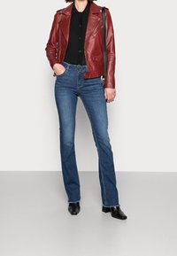 Kobieta w czerwonej skórzanej kurtce, czarnym topie, niebieskich dżinsach z paskiem, czarnych botkach na obcasie i nosząca czarną torbę na ramię, stoi na białym tle.