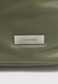 Τσάντα από ύφασμα σε ελιά πράσινο με υφή επιφάνειας και ασημένια μεταλλική ετικέτα που φέρει το όνομα της μάρκας "Calvin Klein". Απλός και λειτουργικός σχεδιασμός.