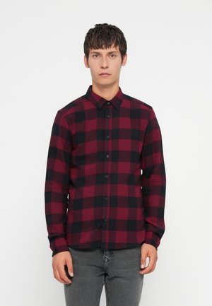Homme portant une chemise à carreaux rouge et noir à manches longues et un pantalon gris, se tenant contre un fond blanc uni.