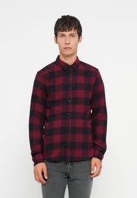 Hombre vestido con una camisa de cuadros roja y negra de manga larga y pantalones grises, de pie frente a un fondo blanco liso.