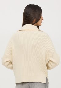 Kremowy, prążkowany sweter z szerokim kołnierzem, opadającymi ramionami i skróconym dołem. Teksturowany materiał i luźny krój.