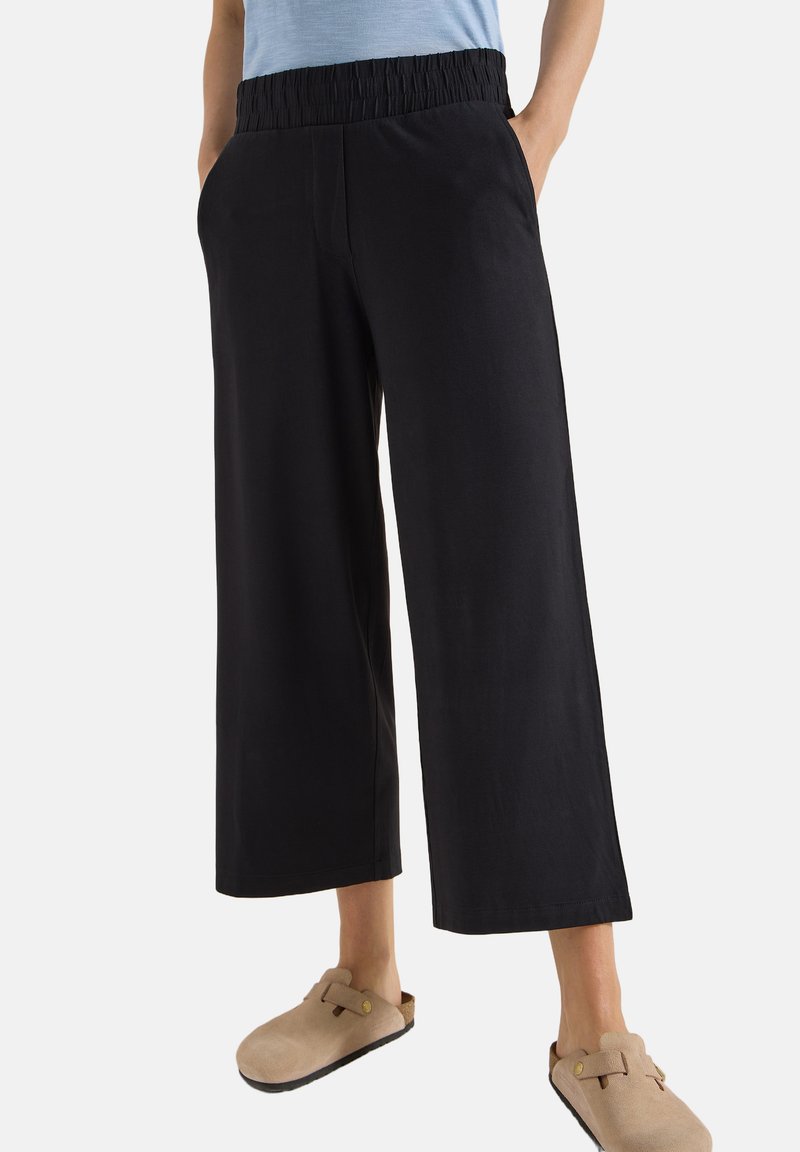Personne portant un pantalon noir large et court avec une taille élastique et des chaussures beiges à enfiler avec des brides à boucle.