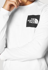 Langärmliges weißes T-Shirt mit einem schwarzen Quadratlogo und dem Text "THE NORTH FACE" sowie einem geschwungenen Designelement auf der oberen linken Brust.