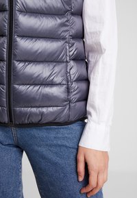 Grå quiltad puffväst med vertikala kanaler och blixtlås i full längd, bärs ovanpå en vit långärmad skjorta och blå jeans.