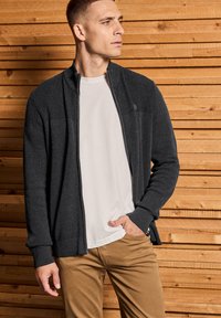 Man i mörkgrå zip-up tröja över vit skjorta och beige byxor, stående mot en vägg av träpanel, tittar åt sidan.