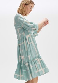 Ein hellblaues, gestuftes Kleid mit weißen geometrischen Mustern, Dreiviertelärmeln und einem weichen, strukturierten Stoff, ideal für den Freizeitlook.