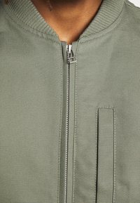 Oliwkowa bomberka z fakturowanej tkaniny, z prążkowanym kołnierzem, z zamkiem błyskawicznym i detalem bocznej kieszeni.