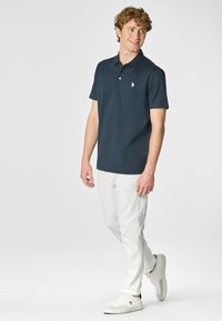 Polo shirt blu navy con maniche corte, colletto e logo sul petto, abbinato a pantaloni bianchi e sneaker bianche.