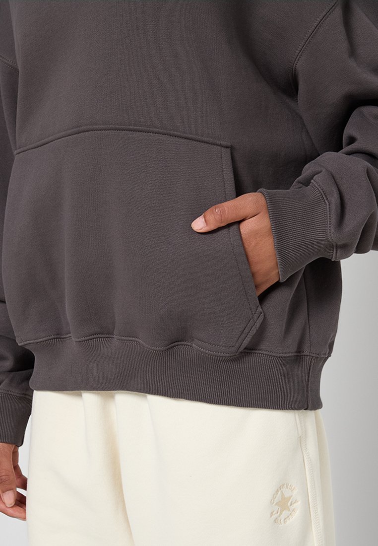 Osoba ubrana w ciemnoszary hoodie z przednią kieszenią oraz jasne kremowe dresy z wyszywanym logo Converse na udzie.
